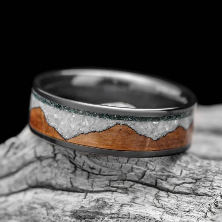 Black titanium wedding band with moss agate, whiskey barrel oak, and pearl inlays, 7mm mens ring, unique design, free engraving, displayed on white sea branch.
