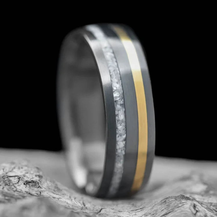 Black titanium wedding band with pearl and 18K yellow gold inlays, featuring free engraving and a brushed domed profile, 7mm mens unique wedding ring displayed on a white sea branch.