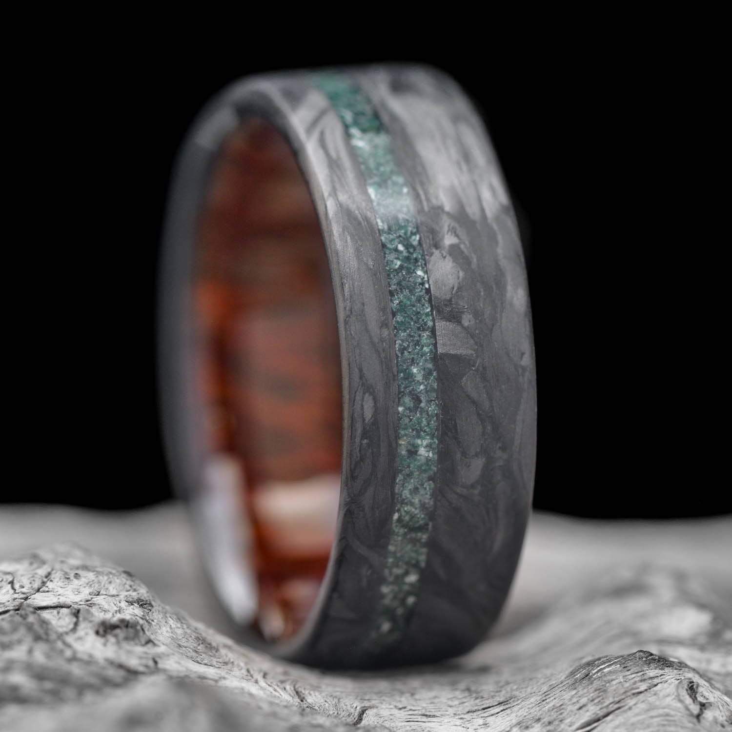 A men's 8mm forged carbon fiber wedding band featuring a vivid green moss agate inlay and a warm leopardwood inner sleeve, offering free engraving, displayed on a white sea branch.
