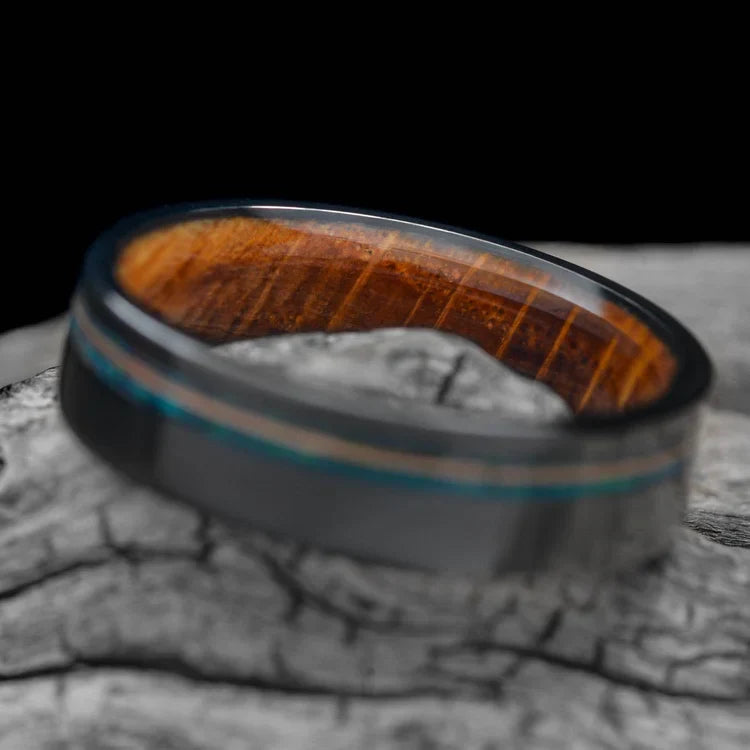 Black titanium wedding band with blue opal and guitar string inlay, featuring a whiskey barrel oak inner sleeve, displayed on a white sea branch, mens 6mm ring with free engraving option.