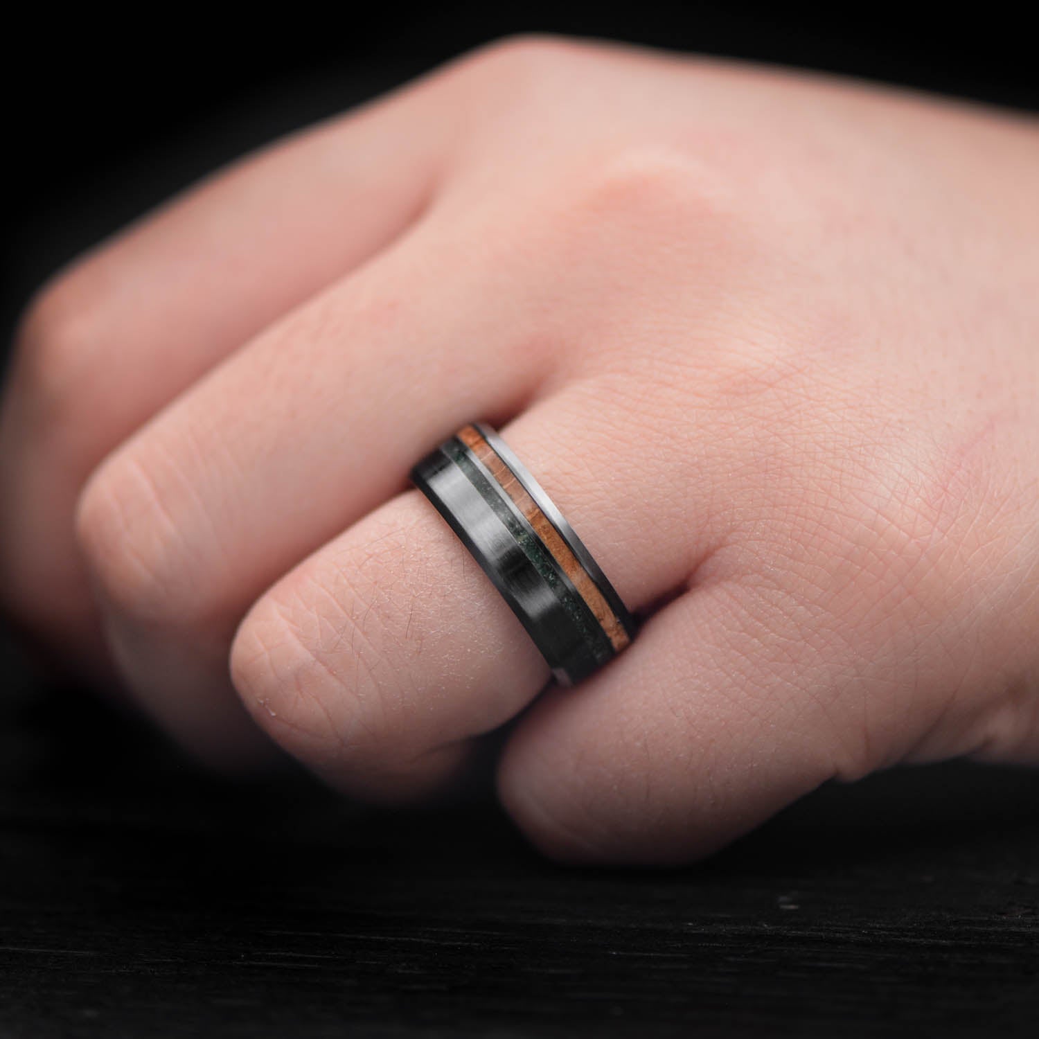 Black titanium wedding band with whiskey barrel oak and moss agate inlays, 8mm mens ring, unique custom design, free engraving, displayed on a white sea branch.