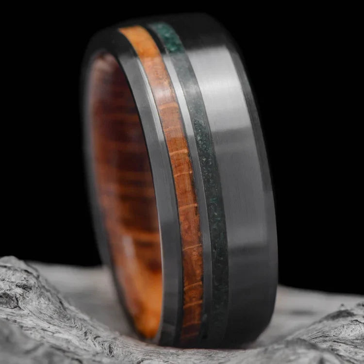 Mens 8mm black titanium wedding band with whiskey barrel oak and moss agate inlays, unique custom design, free engraving, displayed on a white sea branch.