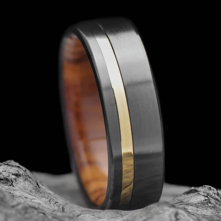Black titanium wedding band with 18K yellow gold inlay and olivewood inner sleeve, 7mm mens ring with brushed flat profile, free engraving, displayed on white sea branch.