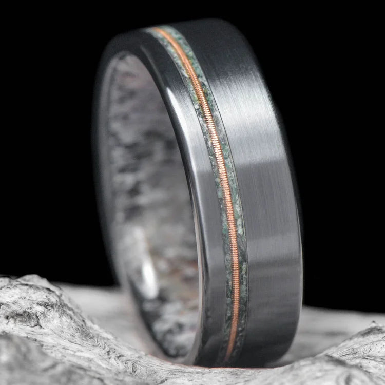 Black titanium wedding ring featuring a moss agate and guitar string inlay with a deer antler inner sleeve, displayed on a white sea branch background; 7mm mens band with free engraving, unique design.