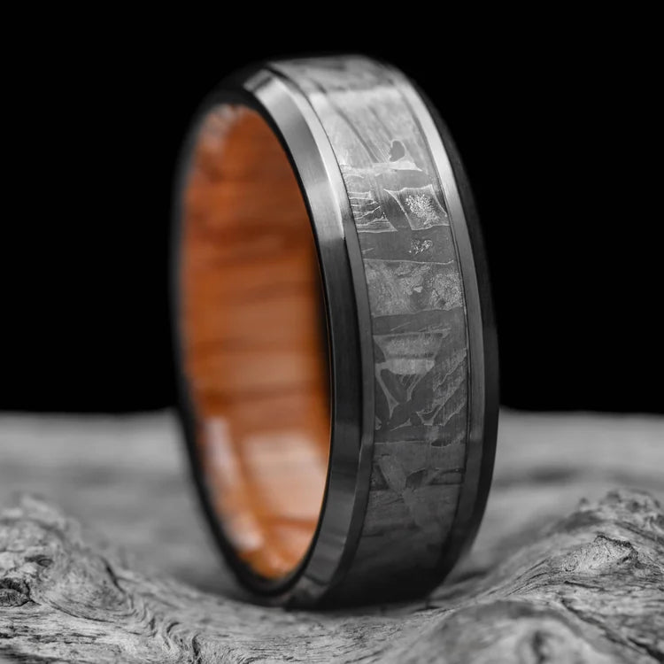 Black titanium wedding band with solid meteorite central inlay and oak inner sleeve, featuring free engraving, 7mm mens brushed beveled ring, displayed on white sea branch.