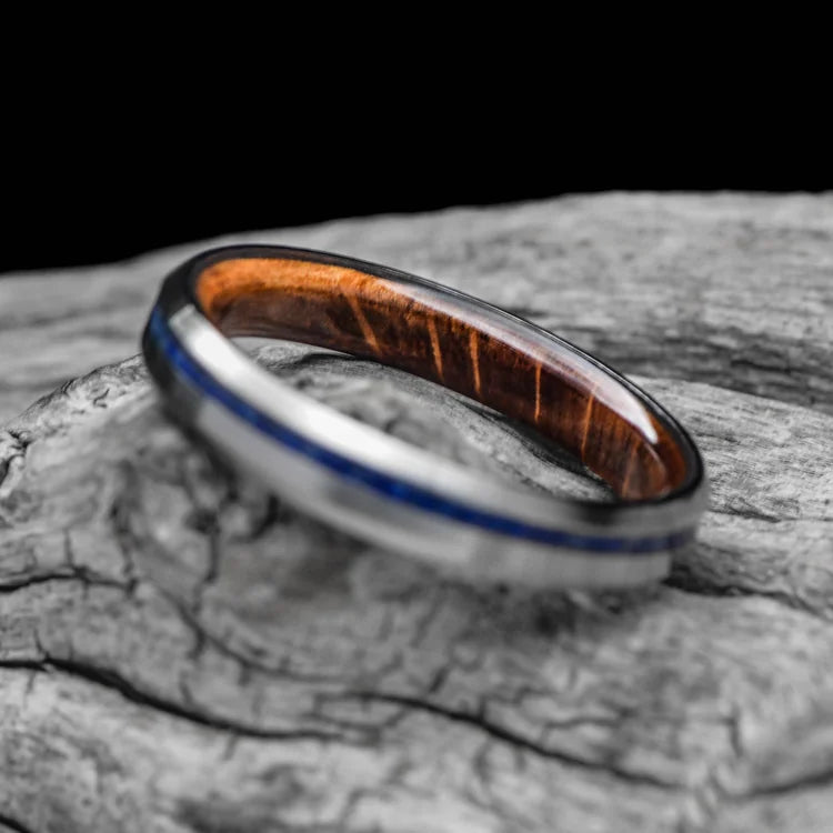 Titanium wedding band with lapis lazuli inlay and whiskey barrel oak sleeve, featuring free engraving, 4mm custom simple ring with brushed bevel profile, displayed on a white sea branch.