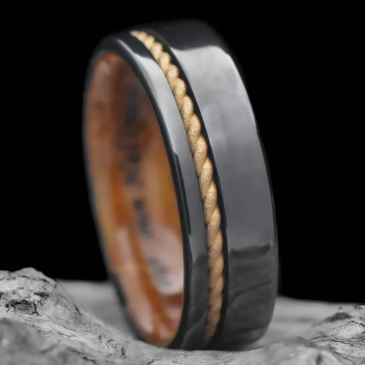 Black titanium wedding band with 10K yellow gold rope and whiskey barrel oak inlay, 7mm mens unique ring, displayed on white sand beach, free engraving available.