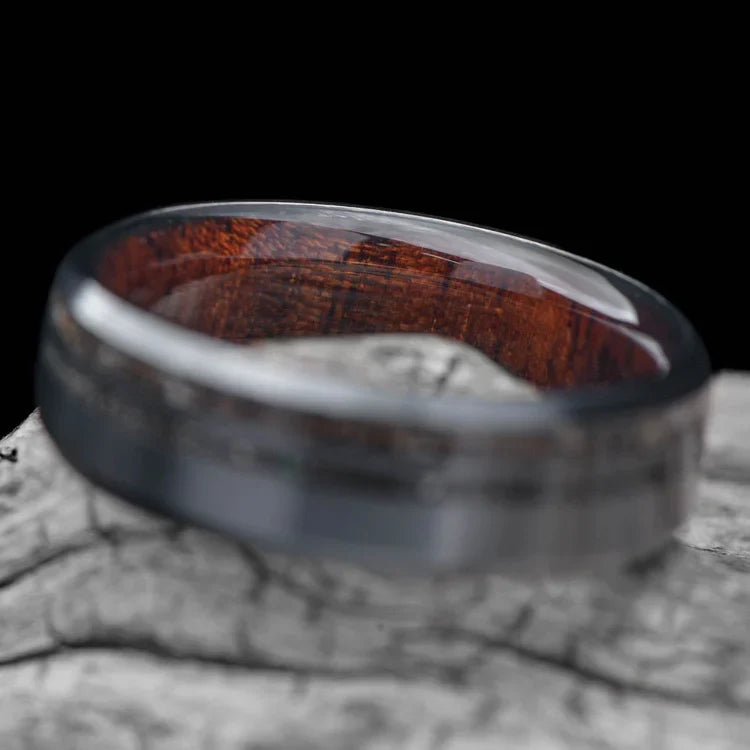 Black titanium wedding band with crushed T-Rex and meteorite inlays, featuring a 7mm width and a unique WW2 rifle inner sleeve, displayed on a white sea branch. Perfect mens ring with free engraving option.