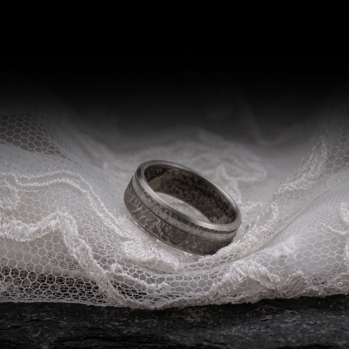 Textured grey wedding band resting on white wedding dress lace over dark slate
