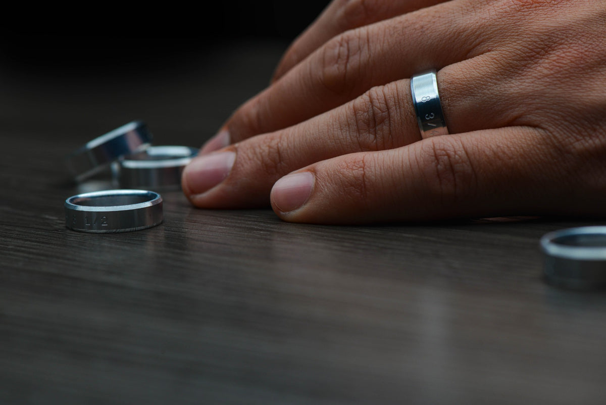 A hand resting on a dark wood table beside three metal Sesto Synergy ring sizers, one worn on a finger